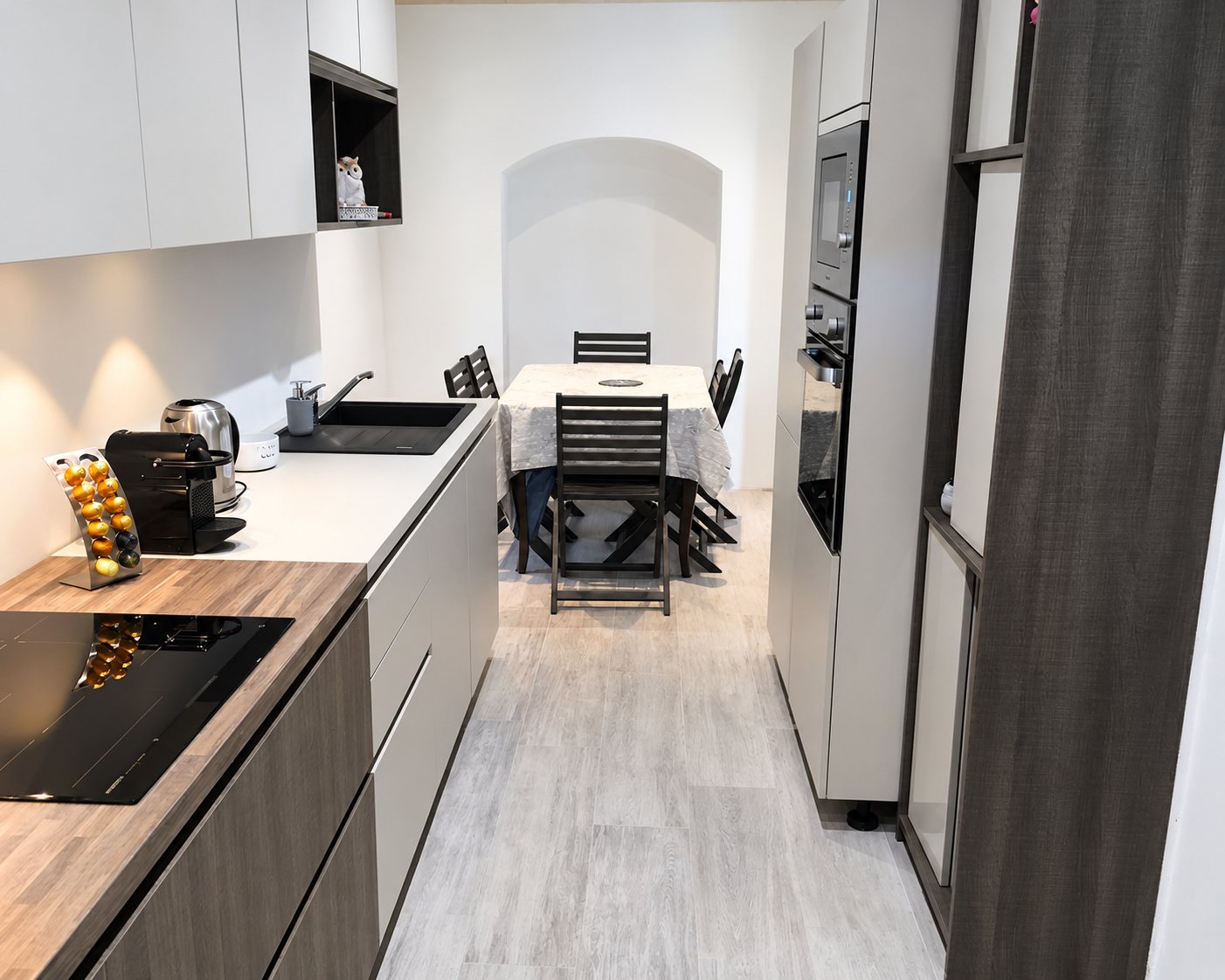 Une cuisine moderne en longueur aux façades grises et claires, avec plaque à induction, four encastré et table à manger au fond.