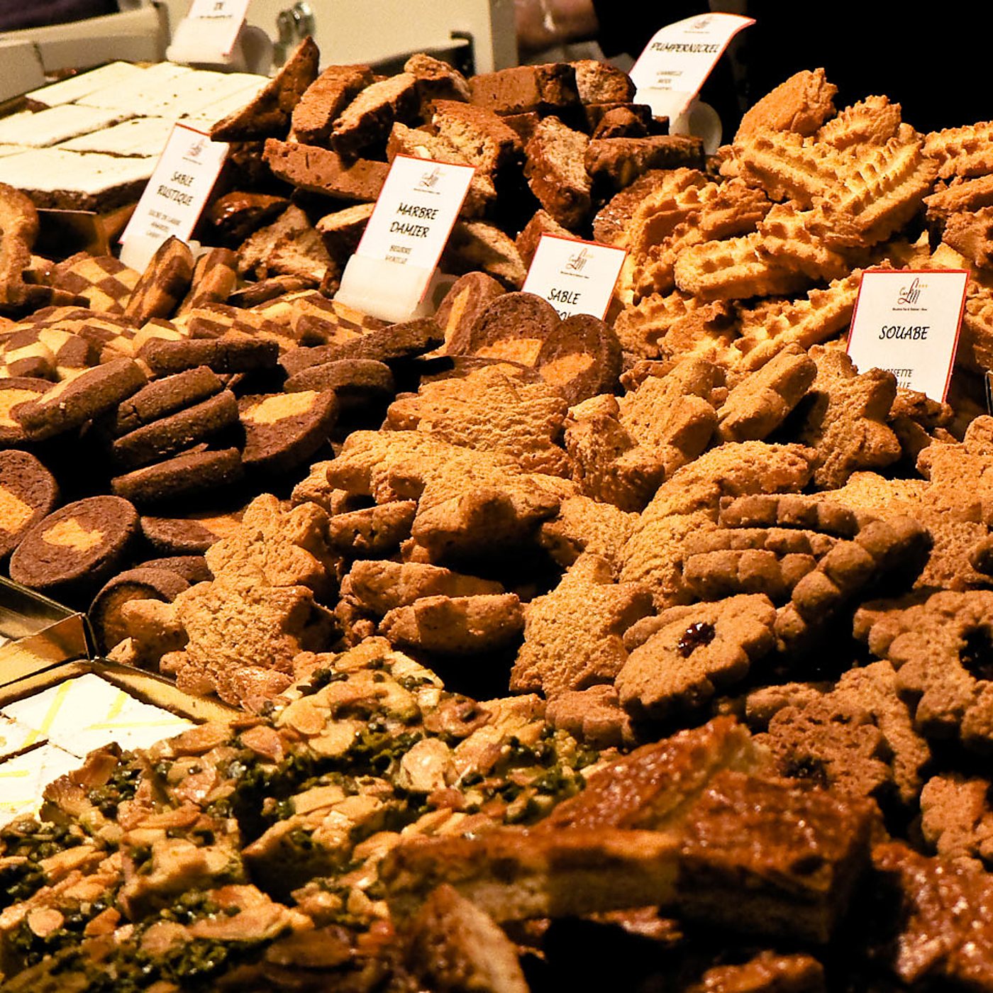 Étal de marché garni de nombreux biscuits, sablés, damiers et gâteaux aux amandes, chacun signalé par une petite étiquette blanche.