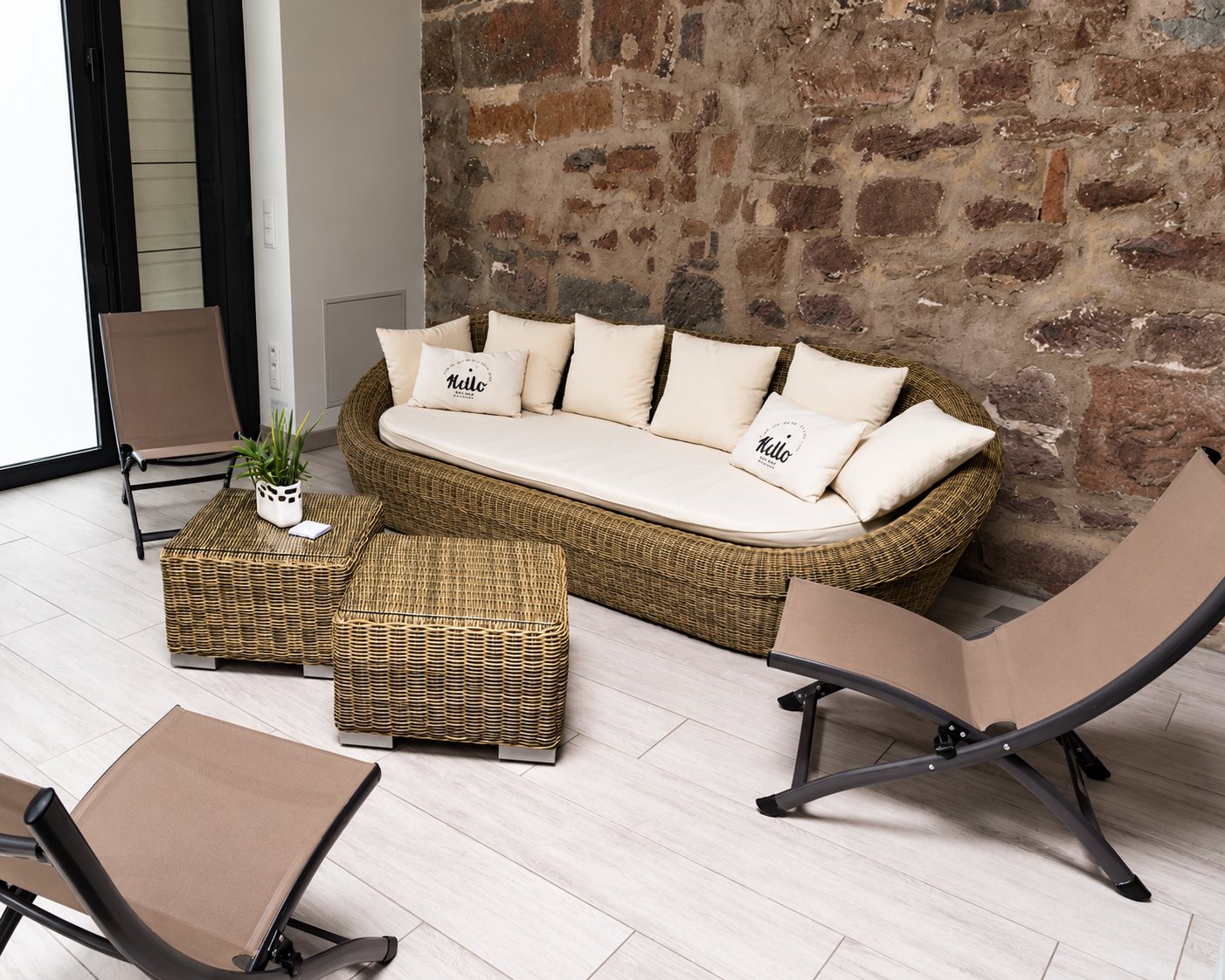 Un coin salon avec un canapé en rotin tressé, des coussins beiges, deux tables basses en osier et des chaises pliantes marron devant un mur en pierre.