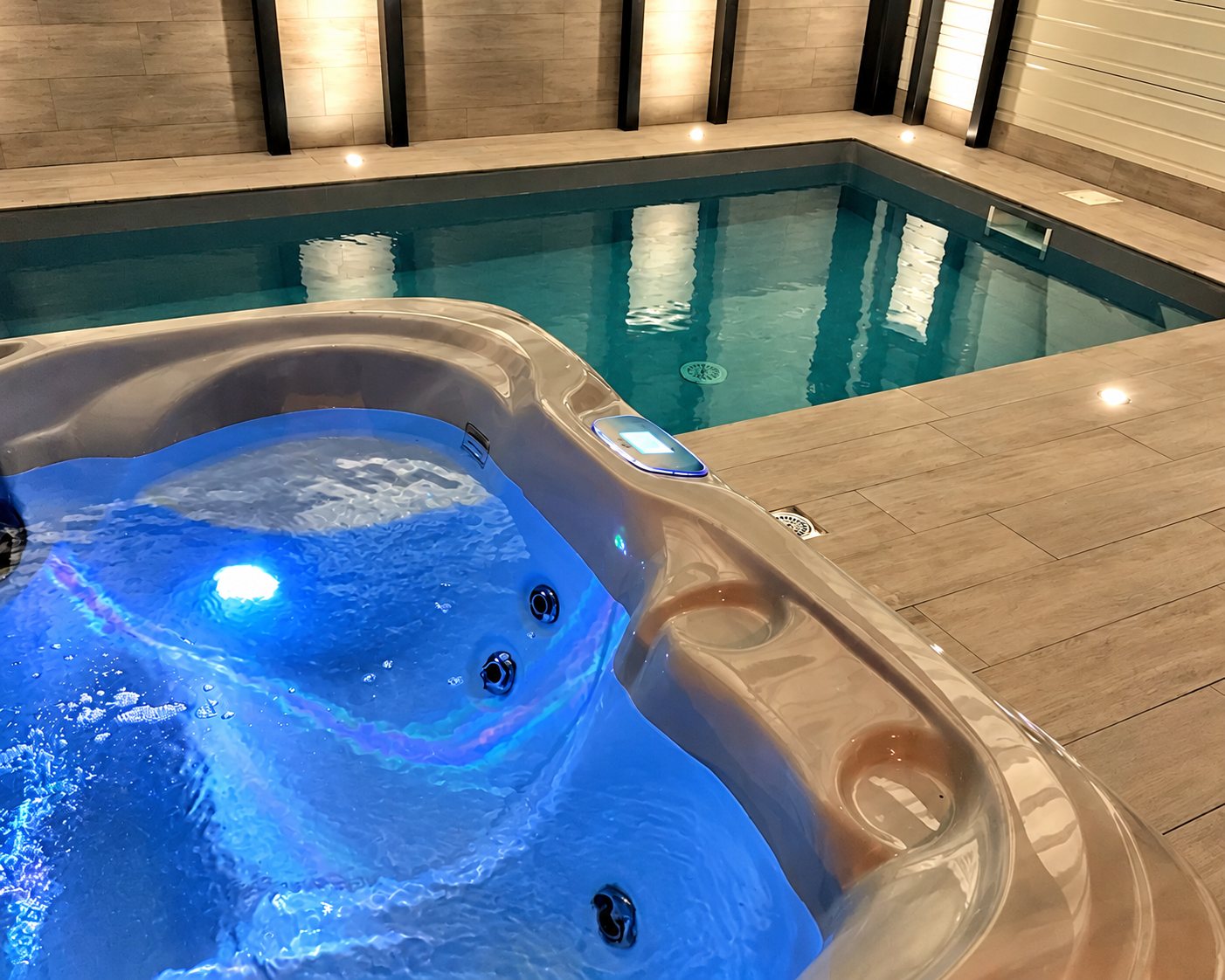 Un spa avec éclairage bleu au premier plan, à côté d'une petite piscine intérieure entourée de carrelage imitation bois.