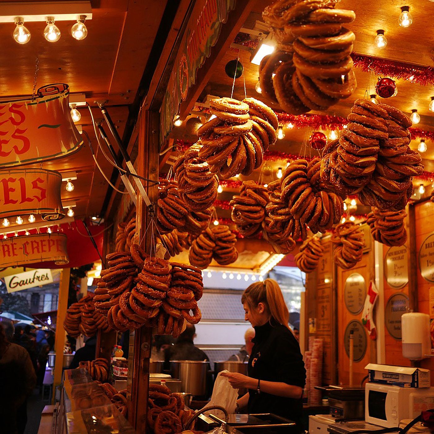 Une vendeuse travaille dans un stand de marché de Noël illuminé, sous des grappes de bretzels dorés suspendus au plafond.