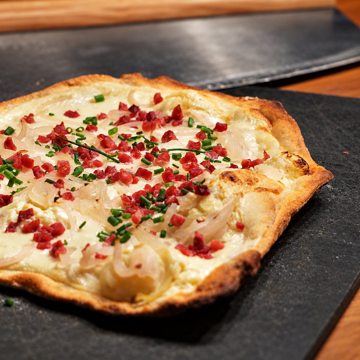 Une tarte flambée alsacienne à la crème, aux oignons, aux lardons et à la ciboulette, posée sur une plaque d'ardoise sombre.