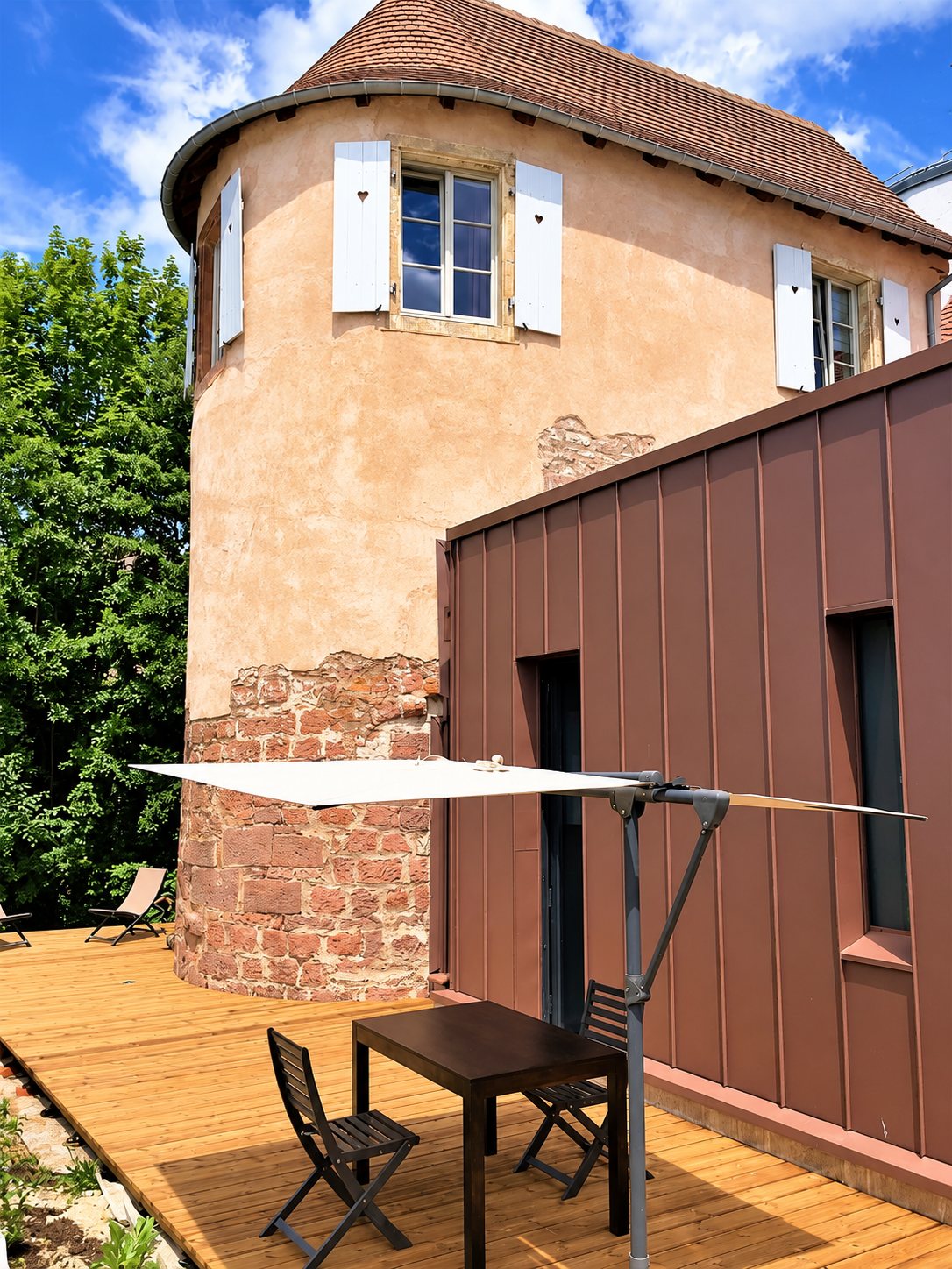 Une ancienne tour en pierre et crépi rose, dotée de volets blancs, jouxte une extension en zinc brun et une terrasse en bois avec table, chaises et voile d'ombrage.