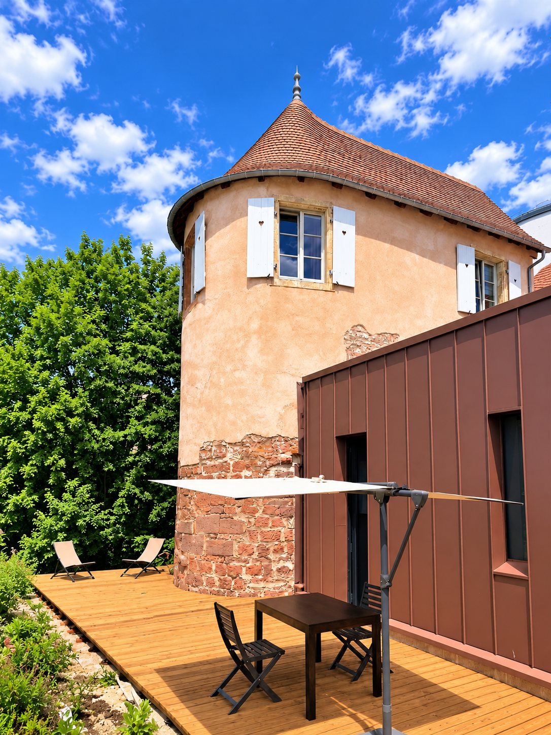 Une ancienne tour en pierre et crépi rose, dotée de volets blancs et d'un toit de tuiles, jouxte une extension en zinc brun et une terrasse en bois avec table, chaises et voile d'ombrage.